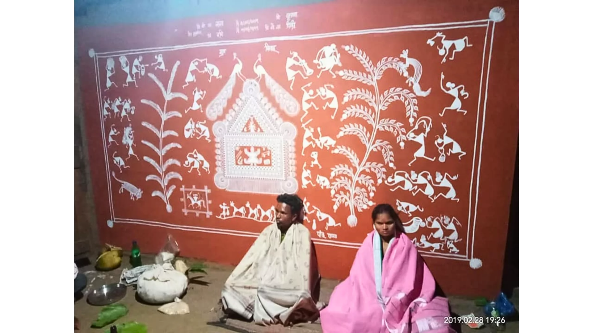 A traditional Warli painting done in white over a red ochre background. Panchasiriya is in the dev chauk in the bottom left of the picture. there is a lagna chauk in the centre.
